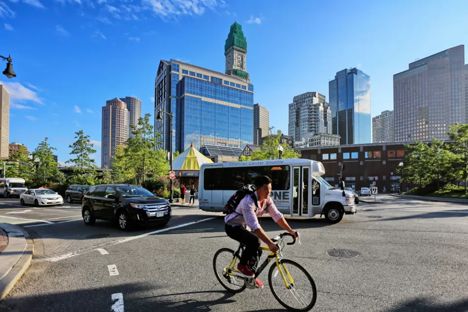 Person on bike wearing backpack with small bus and Boston buildings in background